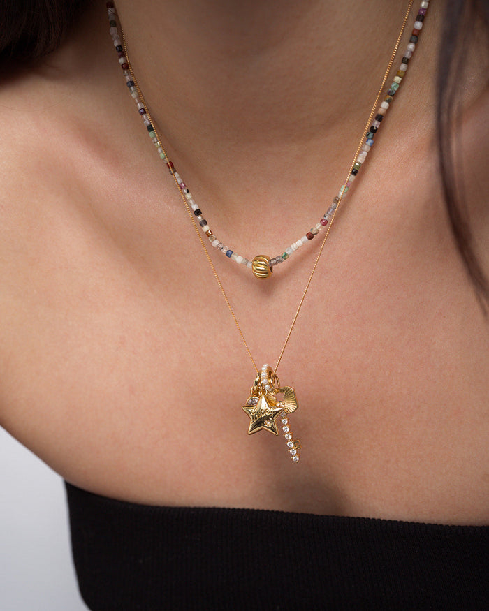 A person wearing two layered necklaces from Mod + Jo: one featuring colorful beads accented with a gold plated bead, the other adorned with the Noah Key Charm alongside gold star-shaped and crystal charms—beautifully displaying these symbolic pieces against bare skin and a black off-the-shoulder top.