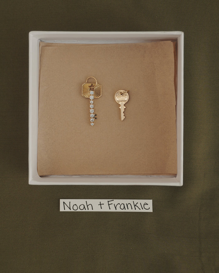 A white box on olive fabric holds two Mod + Jo Noah Key Charms, each gold plated and displayed on brown paper. This symbolic jewelry is accompanied by a handwritten label below that reads Noah + Frankie.