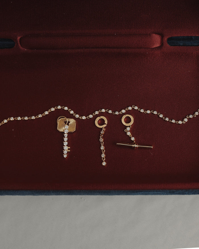 The Mod + Jo Demi Tennis Charm set showcases a dazzling 14k gold-plated chain and matching earrings, both designed with circular tops and delicate dangling accents featuring a sparkling CZ charm, beautifully presented on a deep red velvet backdrop.