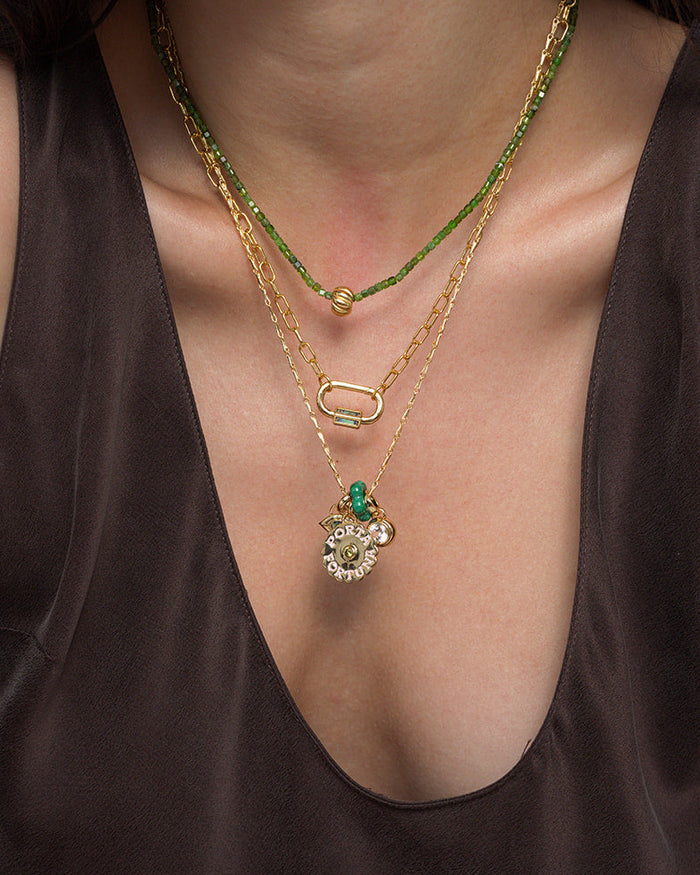 A person wearing Mod + Jo's Nora Lock Carabiner—the luminous, three-layered gold necklace set crafted with 14k plating. One strand is adorned with natural green semi-precious stone beads, another features a refined oval link, and the third showcases a round pendant accented by delicate charms. The look is completed with a deep v-neck brown top.