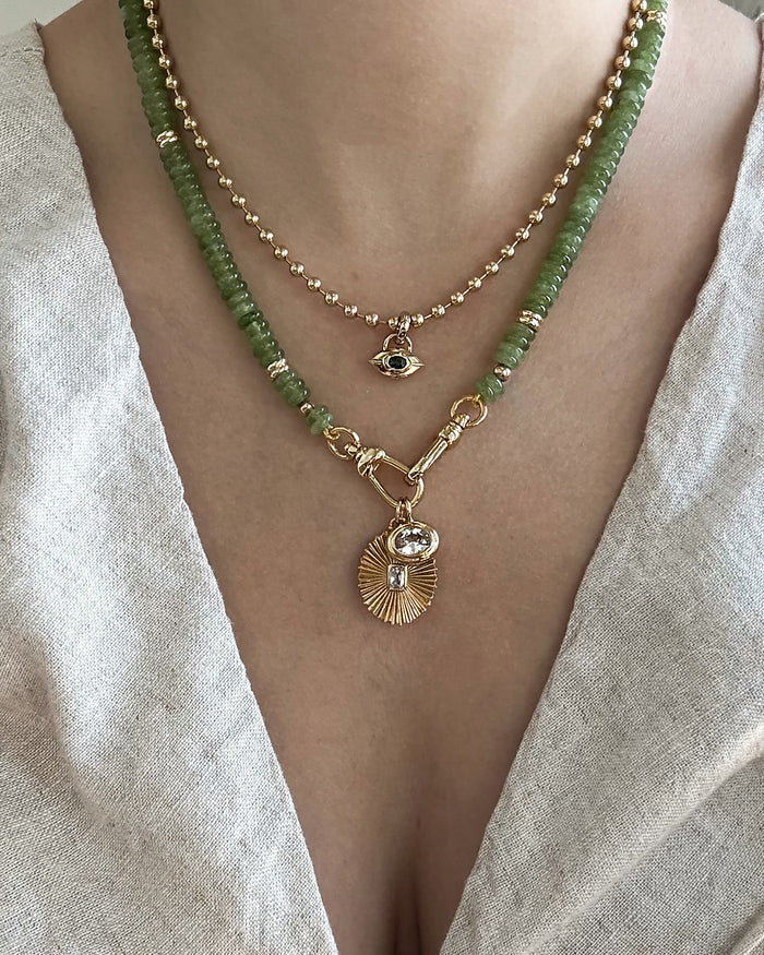 A close-up of a person wearing layered necklaces from Mod + Jo: one features green beads and a gold sunburst protective pendant, while the other is adorned with small gold beads and the Nyx Evil Eye Charm, styled over a light linen V-neck top.