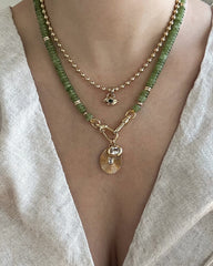 A close-up of a person wearing layered necklaces from Mod + Jo: one features green beads and a gold sunburst protective pendant, while the other is adorned with small gold beads and the Nyx Evil Eye Charm, styled over a light linen V-neck top.