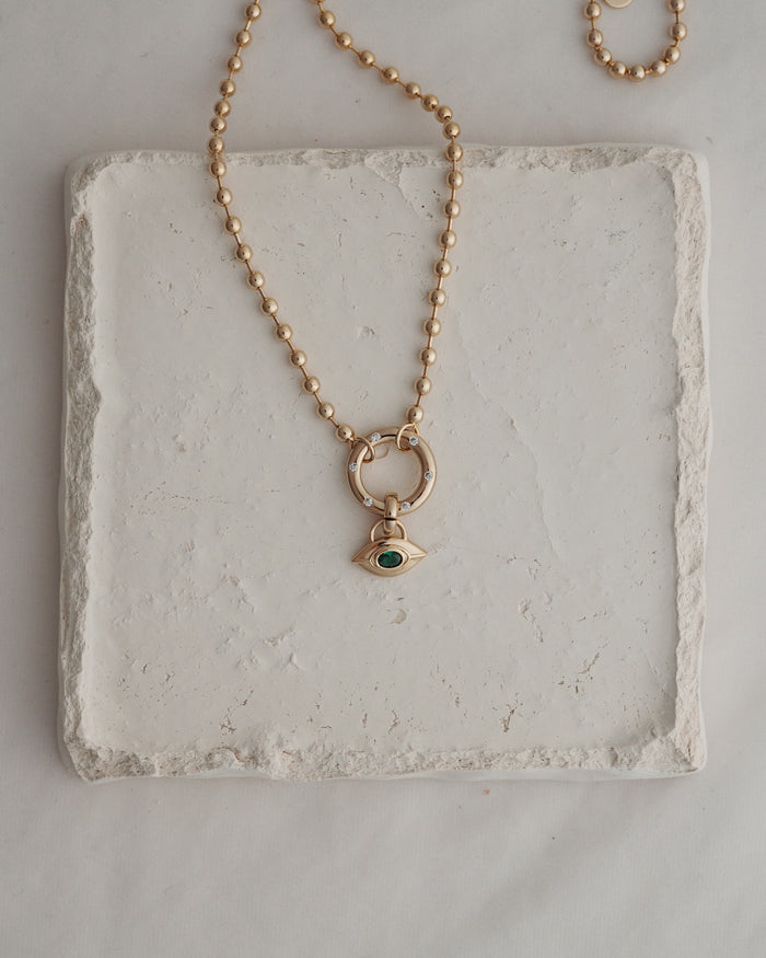 The Nyx Evil Eye Charm necklace by Mod + Jo features a round beaded gold chain and an eye-shaped pendant set with a green gemstone, artfully displayed on a textured white square tile—an elegantly crafted piece of protective jewelry that exudes the subtle allure of the evil eye.
