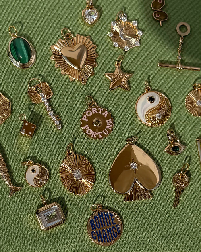Mod + Jo gold and jeweled charms—including hearts, stars, yin-yangs, a protective pendant, and the Nyx Evil Eye Charm—are displayed on a green textured fabric background.
