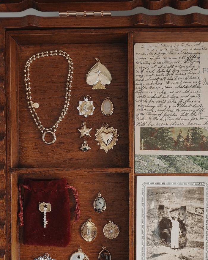 A wooden jewelry box contains gold charms, a bracelet, a protective Mod + Jo Nyx Evil Eye Charm pendant, a red velvet pouch holding a key, vintage postcards, and a black-and-white photograph of a person standing outside against a stone wall.