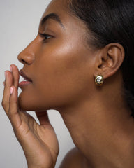 A woman with smooth, glowing skin is shown in profile, gently touching her face. She wears the Penn Gemstone Hoops by Mod + Jo—gold-plated over brass, nickel and lead-free, featuring sparkling CZ accents and a clear gemstone. The background is neutral and uncluttered.