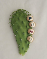 A green cactus pad displays four Mod + Jo Penn Gemstone Hoops on its right edge, each hoop adorned with different gemstones and sparkling CZ accents to resemble vibrant fingernails. The background features a soft, neutral fabric.
