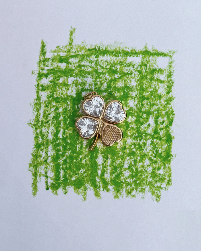 A gold Perry Four-Leaf Clover Locket Charm by Mod + Jo, featuring three leaves adorned with clear gemstones, rests on a green crayon square drawn on white paper, evoking the whimsy and elegance of cherished keepsakes.
