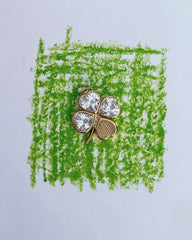 A gold Perry Four-Leaf Clover Locket Charm by Mod + Jo, featuring three leaves adorned with clear gemstones, rests on a green crayon square drawn on white paper, evoking the whimsy and elegance of cherished keepsakes.