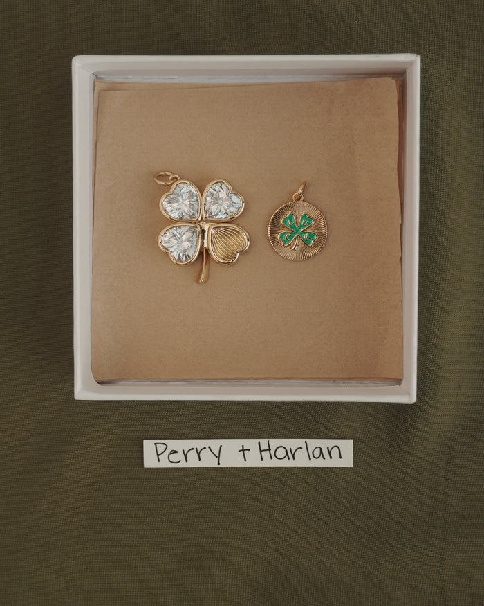 A white box displays the Perry Four-Leaf Clover Locket Charm by Mod + Jo—a gold locket in the shape of a four-leaf clover, its leaves shimmering with heart-cut CZ stones, paired with a small gold medallion adorned with a green clover. The strip below reads "Perry + Harlan.