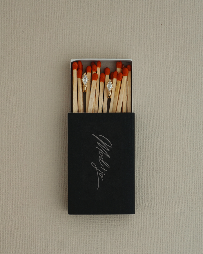 A black matchbox with “Marlsto” written on it, filled with matches, displays two matches adorned with sparkling Phoebe Kite Huggies by Mod + Jo—each shining like dazzling cubic zirconia earrings—set against a neutral textured background.