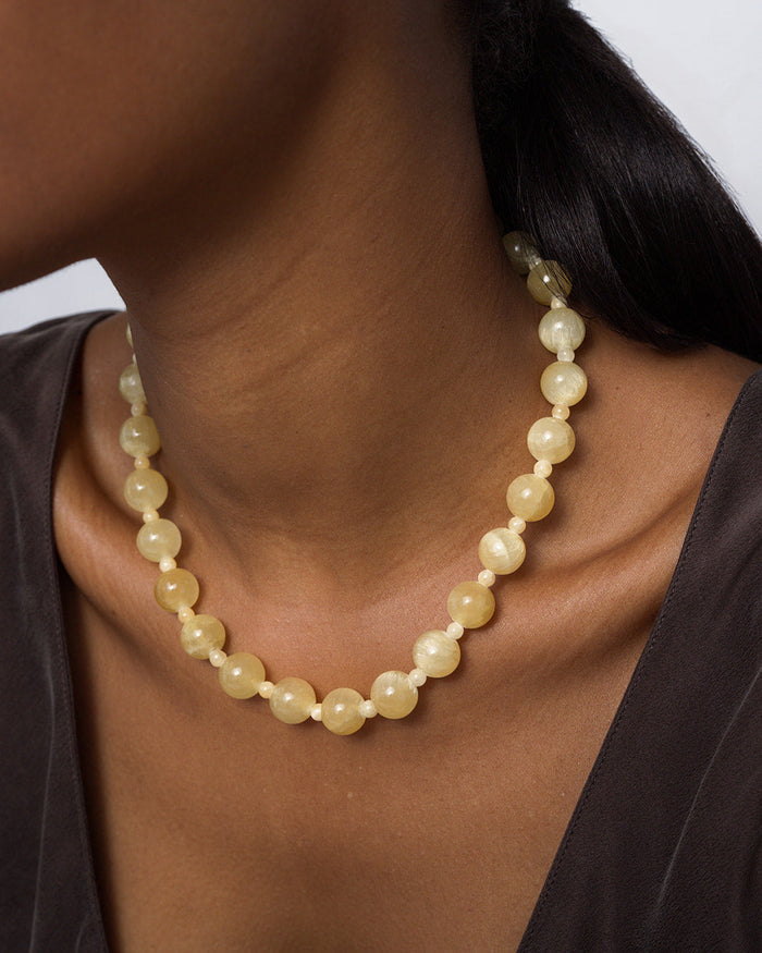 A person with dark, pulled-back hair wears a dark V-neck top and the Mod + Jo Portia Gemstone Necklace, featuring round, pale yellow beads that rest comfortably around the neck.