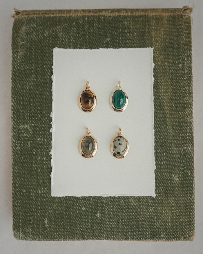 Four Rory Gemstone Charms by Mod + Jo, each featuring a unique oval gemstone with 14k gold over brass edges, are arranged on a sheet of white paper centered against a textured olive green square background. Each charm showcases a different stone—including the striking Dalmatian Jasper—and is thoughtfully crafted for an elegant touch.
