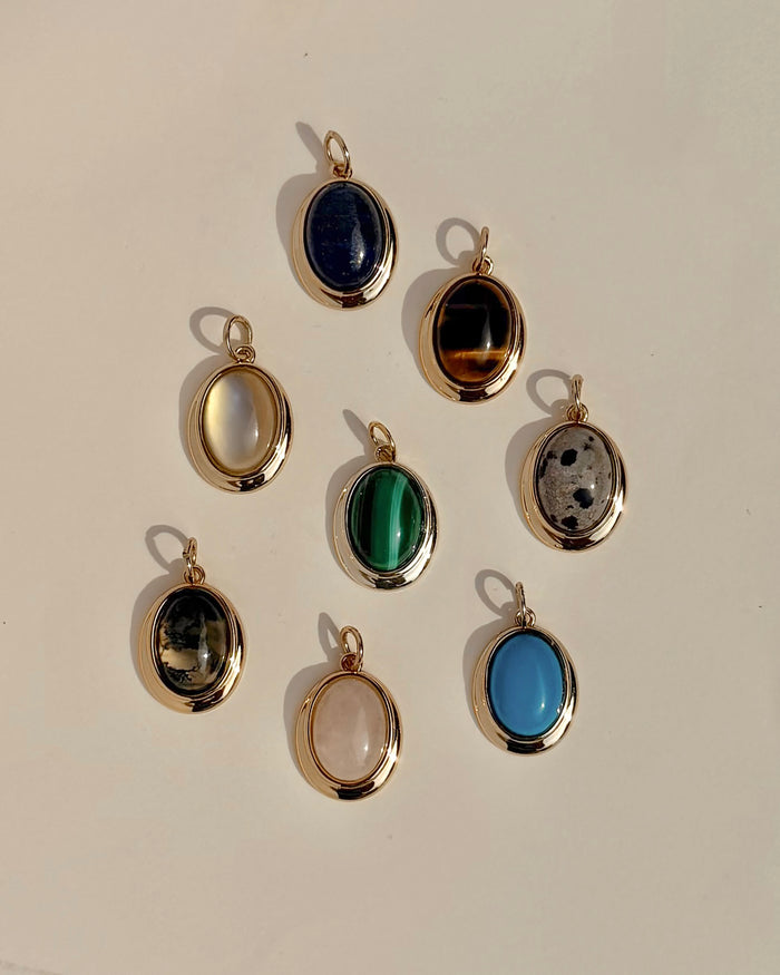 Eight exquisite Rory Gemstone Charms by Mod + Jo, each framed in 14k gold, are elegantly arranged on a light surface. Every charm showcases a distinct gemstone hue—serene blue, vibrant green, mysterious black, luminous white, gentle pink Rose Quartz symbolizing self-love, and uniquely patterned multicolored stones.