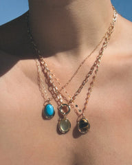 A person wears three Mod + Jo Rory Gemstone Charm gold chain necklaces, each featuring a unique gemstone pendant: a turquoise oval stone, a translucent oval stone, and a brown-and-black striped stone, all set in gold. The jewelry is displayed against bare skin with a blue sky in the background.