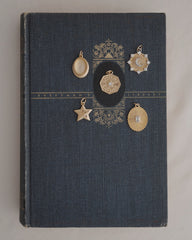 A dark blue vintage book with gold detailing lies on a white surface. Five Rory Gemstone Charms by Mod + Jo, each featuring unique shapes, intricate designs, and shimmering Moonstone accents in gold and silver tones, are arranged on the book’s cover.