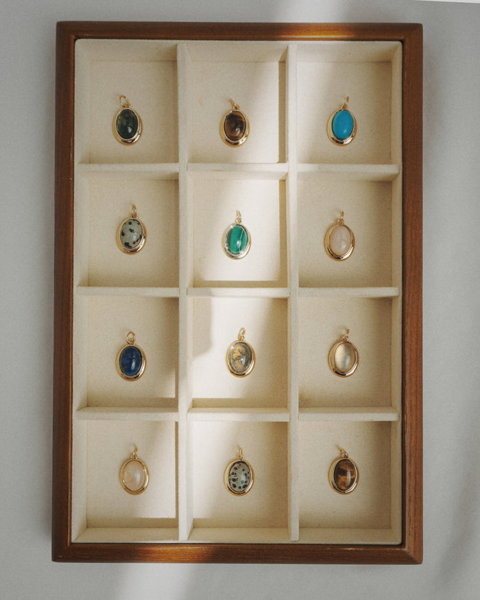 A wooden display case featuring twelve compartments arranged in a 3x4 grid, each showcasing a Mod + Jo Rory Gemstone Charm—a round pendant with a unique colored stone such as exquisite lapis lazuli—beautifully illuminated by natural sunlight.
