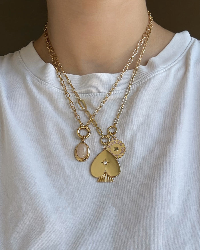 A person wearing a white shirt with two layered 14k gold chain necklaces from Mod + Jo, each adorned with distinctive pendants: a small oval, a spade with a star, and the Rory Gemstone Charm—a round sun-shaped piece that embodies self-love.