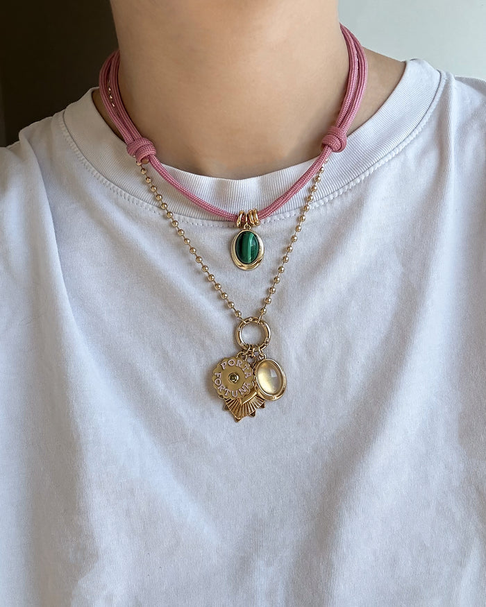 A person wearing a white shirt layered with two necklaces: a pink cord featuring Mod + Jo’s Rory Gemstone Charm—a green oval pendant that shimmers like moonstone—and a gold chain adorned with round gold charms and a small pearl-like locket.