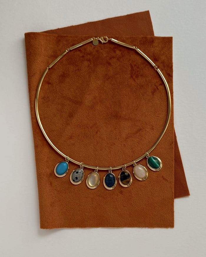 The Mod + Jo Rory Gemstone Charm necklace, featuring seven colorful gemstone pendants and inspired by lapis lazuli jewelry, is displayed on brown suede fabric against a white background.