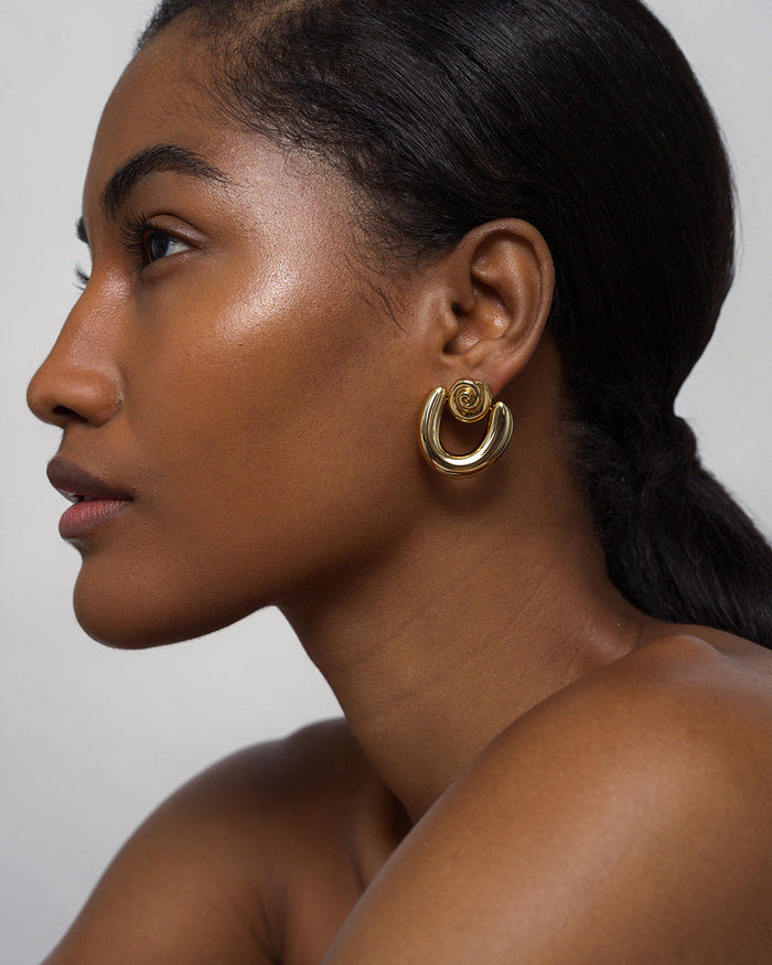 A woman with glowing skin and long dark hair pulled back wears Mod + Jo Sabine Statement Earrings—large, shiny gold hoops with a circular design. She appears in profile against a light, neutral background.