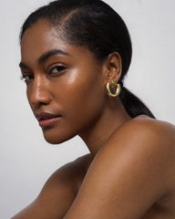 A woman with smooth, glowing skin and sleek dark hair pulled back models the Mod + Jo Sabine Statement Earrings in chunky gold, looking confidently at the camera against a plain light background.