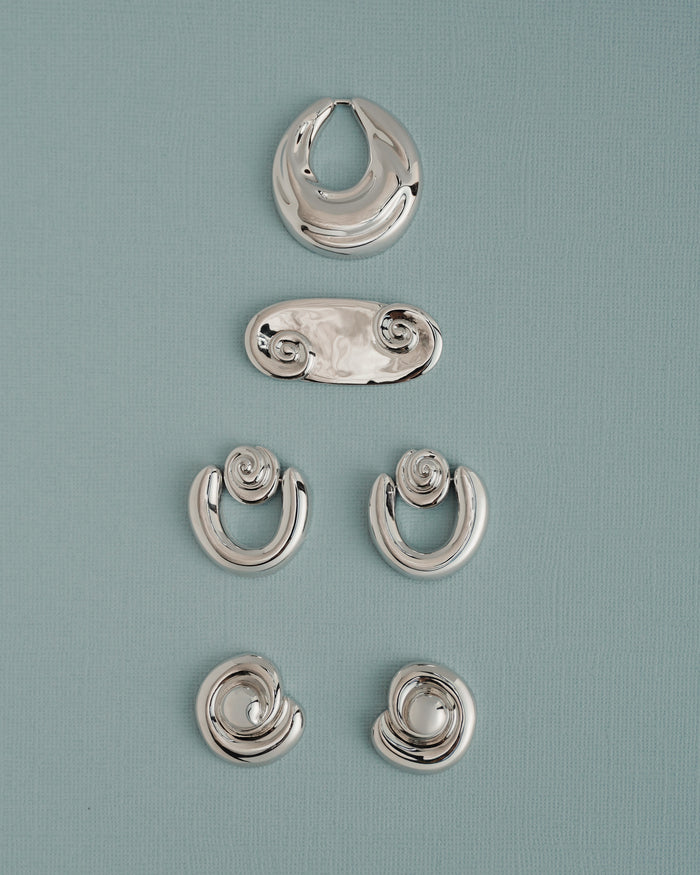 Five pairs of Sabine Statement Earrings by Mod + Jo, featuring shiny silver in abstract and spiral shapes, are displayed vertically on a light blue textured background.