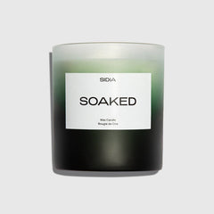 A black and green SOAKED Candle by SIDIA, housed in a frosted glass container with a white label, sits against a plain light gray background. Its subtle notes of juniper and jasmine evoke the refreshing scent of wild herbs.