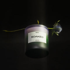 A black SOAKED Candle by SIDIA is displayed against a dark background, accompanied by a spiky purple thistle flower and subtle notes of jasmine behind the candle.