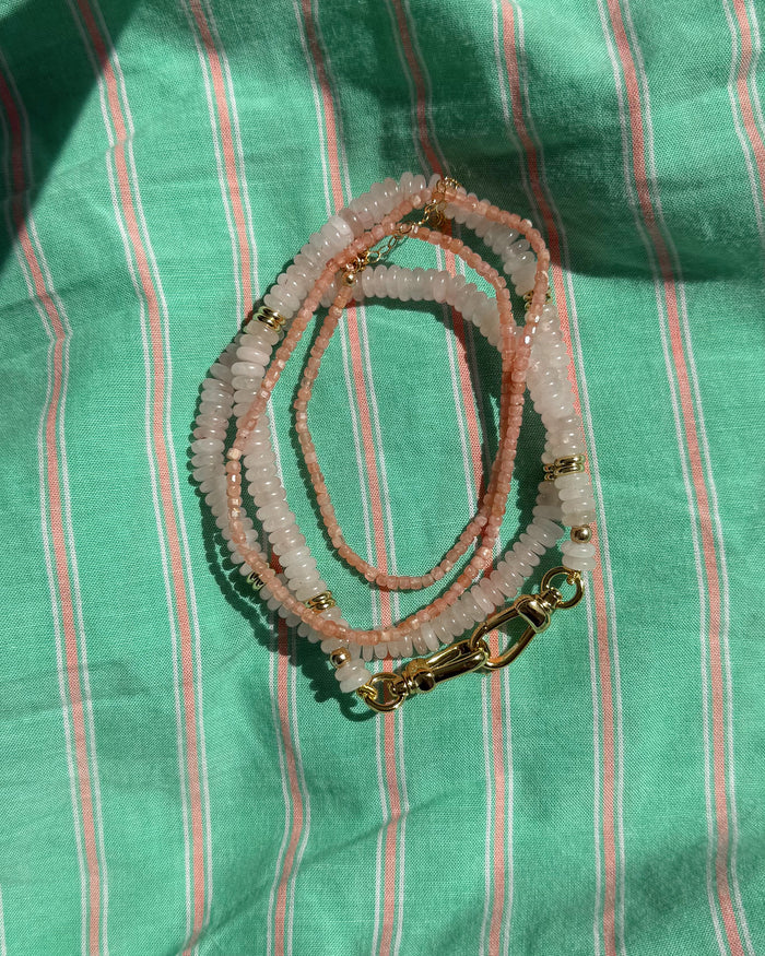The Sonia Gemstone Necklace by Mod + Jo features pink and white genuine gemstone beads with gold clasps, coiled on green striped fabric and shown in natural sunlight—a beautiful piece of handmade jewelry.