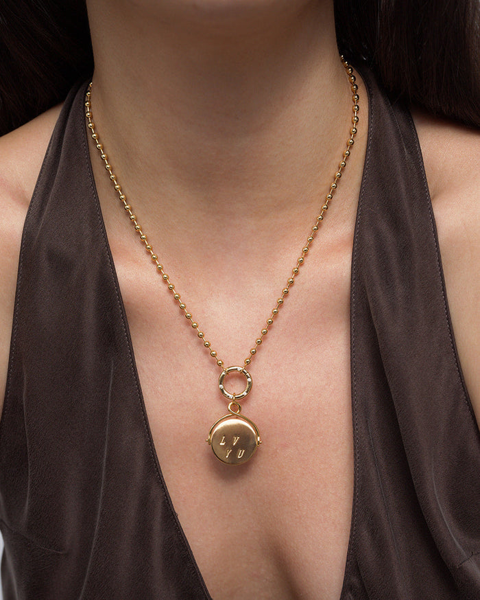 A person wearing a brown satin top and the Mod + Jo Bella "I Love You" Spinner Charm—a 14k gold necklace featuring a round pendant engraved with “LYU”—on a ball chain resting at the center of the chest.