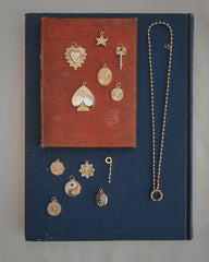 The Soren Ball Charm Clip Necklace by Mod + Jo is displayed on two stacked books, one red and one blue, adorned with gold charms in various shapes such as a heart, star, key, sun, yin-yang, and more—all arranged neatly for viewing.