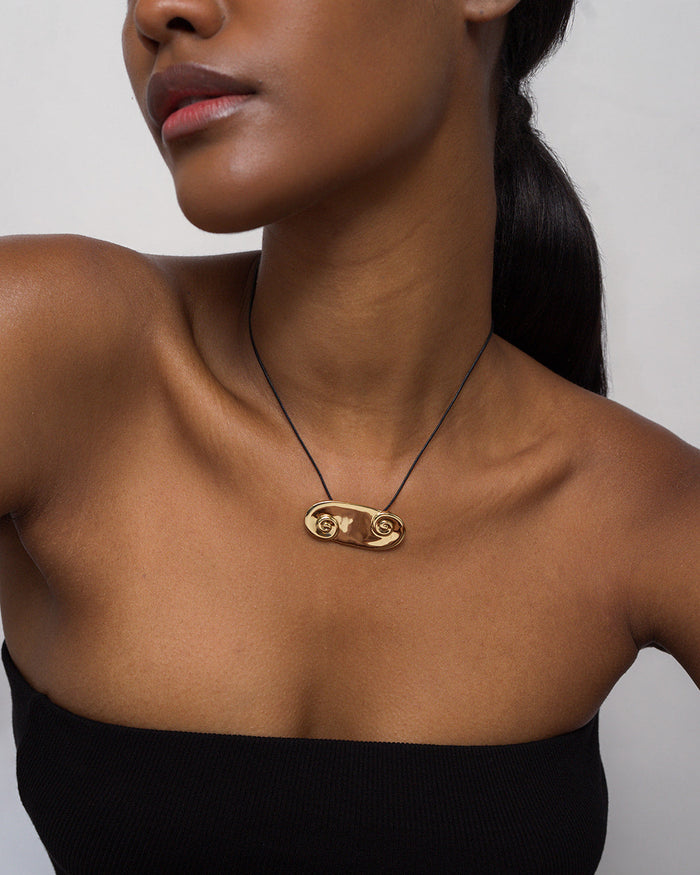 A woman wears a black strapless top and the Mod + Jo Suna Spiral Cord Necklace in 14k gold, featuring an artistic design and an adjustable bolo. Her hair is tied back against a plain, light-colored background.