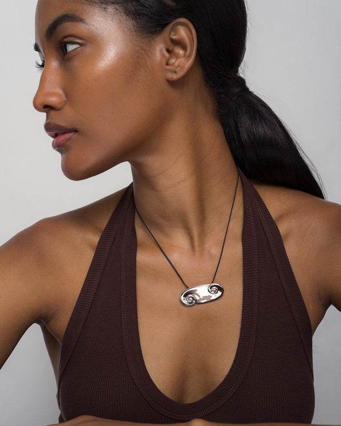 A woman with smooth, glowing skin and dark, straight hair tied back wears a brown halter top and the Mod + Jo Suna Spiral Cord Necklace—a striking double-spiral pendant on an adjustable cord—while looking to the side against a gray background.