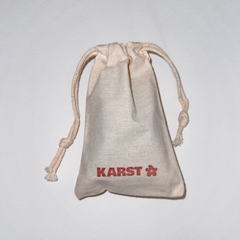 A small beige drawstring pouch with "Karst Essentials" and a red flower symbol printed on the front, perfect for storing your SuperClip Hair Claw — Buttermilk. Photographed against a plain white background.