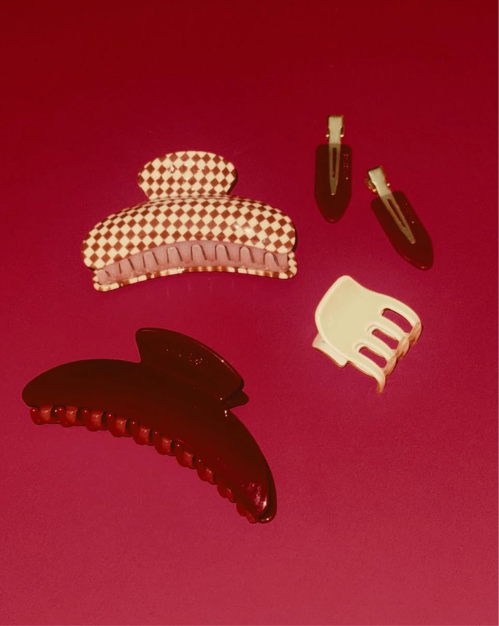 Four SuperClip Hair Claws on a red surface: one in red cellulose acetate, one in white, one checkered white and red, plus two small dark claws with metallic details.