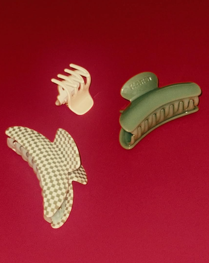 Three SuperClip Hair Claws on a red surface: one small cream, one large green, and one large durable beige and white checkered claw clip.