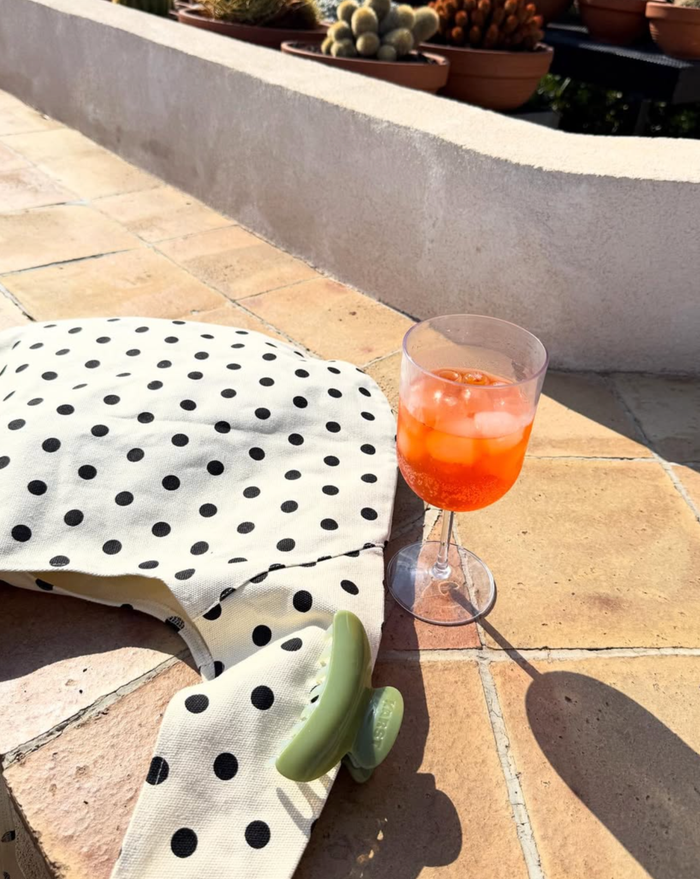A polka dot shirt, a green SuperClip Hair Claw, and an orange iced drink sit on sunlit tiles, with potted plants and cacti in the background.