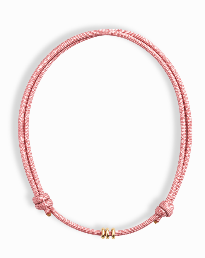 The Tommy Cord Necklace by Mod + Jo features a pink paracord with three gold-plated spacers at the front and adjustable side knots, shown against a white background.