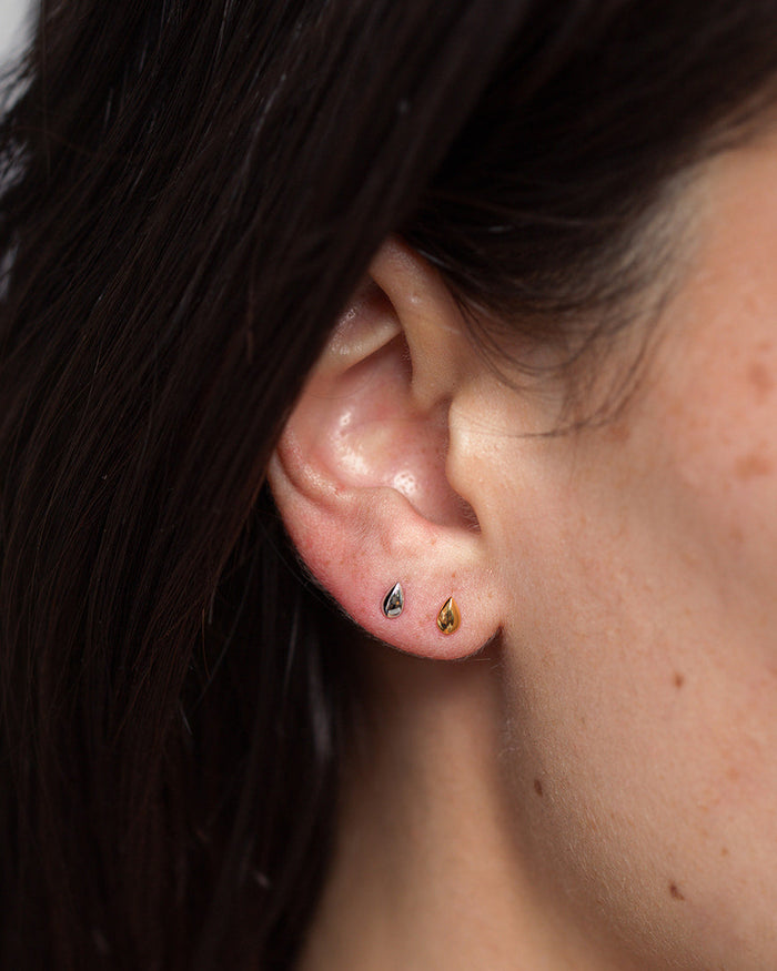 A close-up of a person’s ear features the Uma Teardrop Studs by Mod + Jo—one in gold and one in silver—worn in the earlobe. With dark hair tucked behind the ear, these elegant teardrop-shaped earrings are perfectly showcased, making them an effortless choice for everyday jewelry.