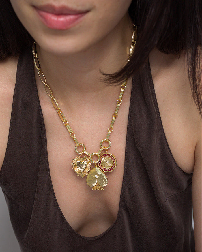 A woman, shown from the shoulders down in a dark brown top, wears the Mod + Jo Veya Spade Charm—an elegant 14k gold necklace featuring three large charms: two heart-shaped pendants, a circular pendant, and a sophisticated gold spade charm accented by a starburst crystal.