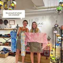 Two women wearing aprons proudly display handmade marbled fabric in colorful art studio with mosaic lamps and ceramics
